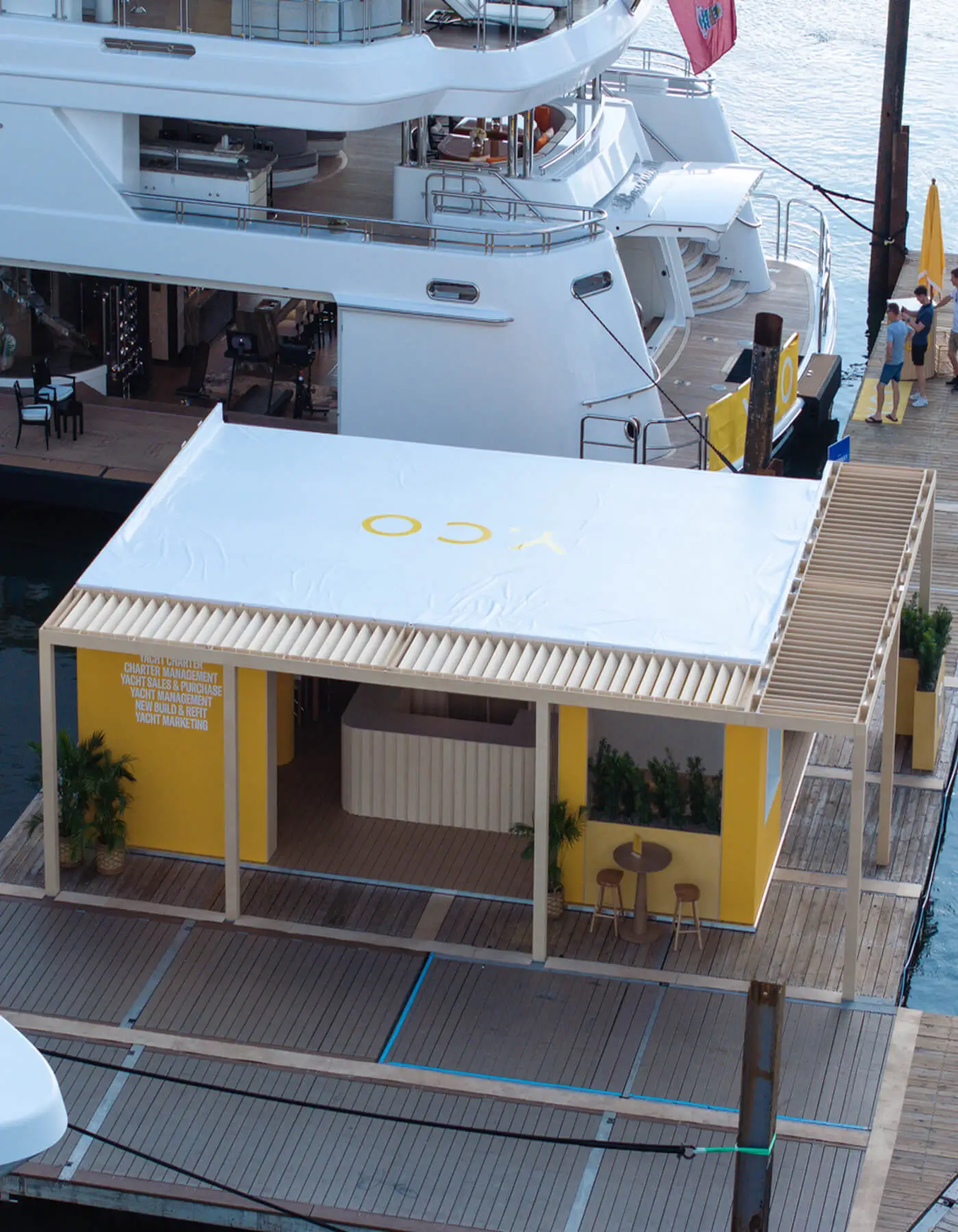 Aerial high-angle view of the modular Y.CO Clubhouse exhibition stand designed by Studio Moravová, built on a floating wooden pontoon dock right next to luxury superyachts at a marina event.