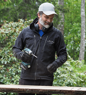 Man in black jacket and gray cap holding a power drill outdoors near wood and greenery.
