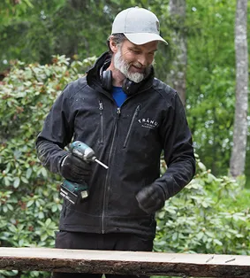 Man in black jacket and gray cap holding a power drill outdoors near wood and greenery.