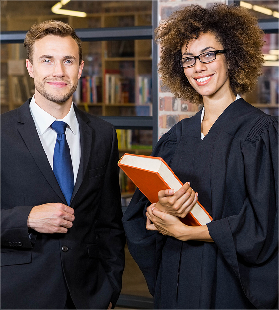 Smiling man in suit and woman in judge robe holding book standing indoors with shelves behind.