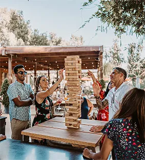 Group of adults playing giant Jenga outside around a wooden table with trees and lights in background.