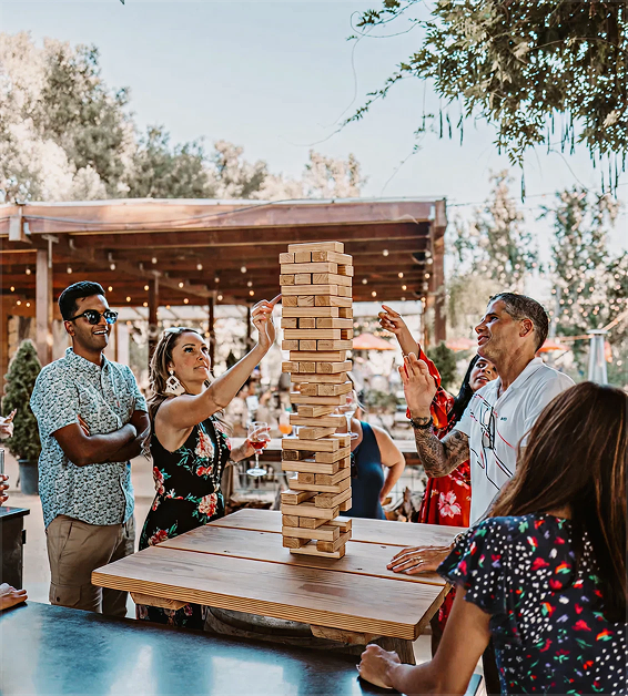 Group of adults playing giant Jenga outside around a wooden table with trees and lights in background.