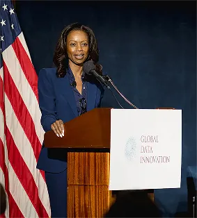 Woman in blue suit speaking at a podium with a Global Data Innovation banner and U.S. flag behind her.