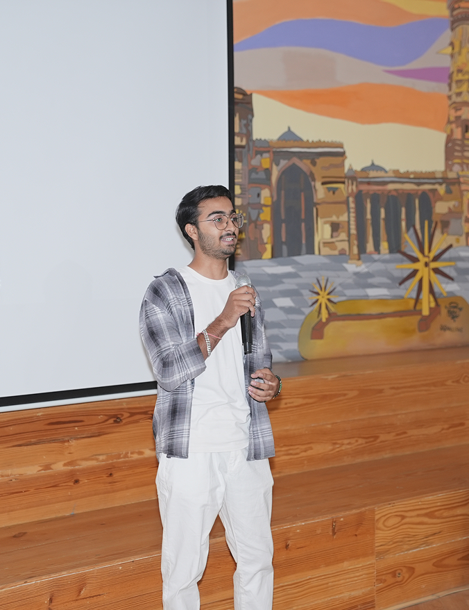 Young man in glasses holding a microphone, speaking in front of a screen and mural in a wooden interior.