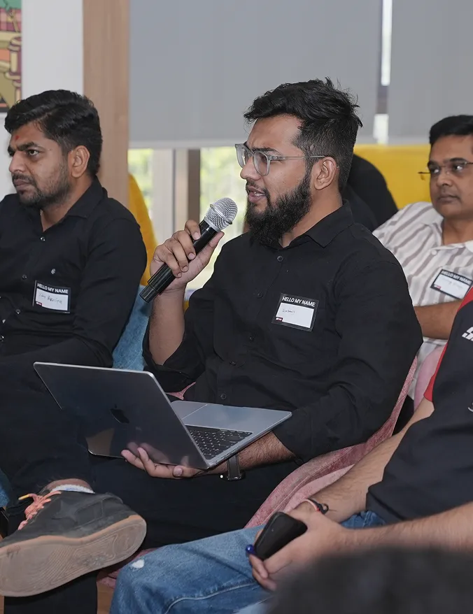 Man holding microphone and laptop speaking in a group with others seated nearby.