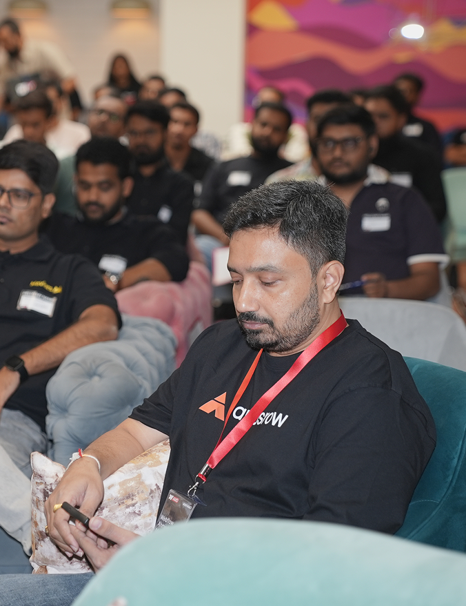 Man wearing a black t-shirt with a red lanyard uses phone in a seated audience at an indoor event.