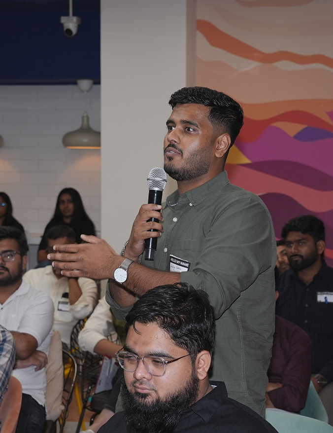 Man in green shirt speaking into microphone during a seated group event with colorful wall art.
