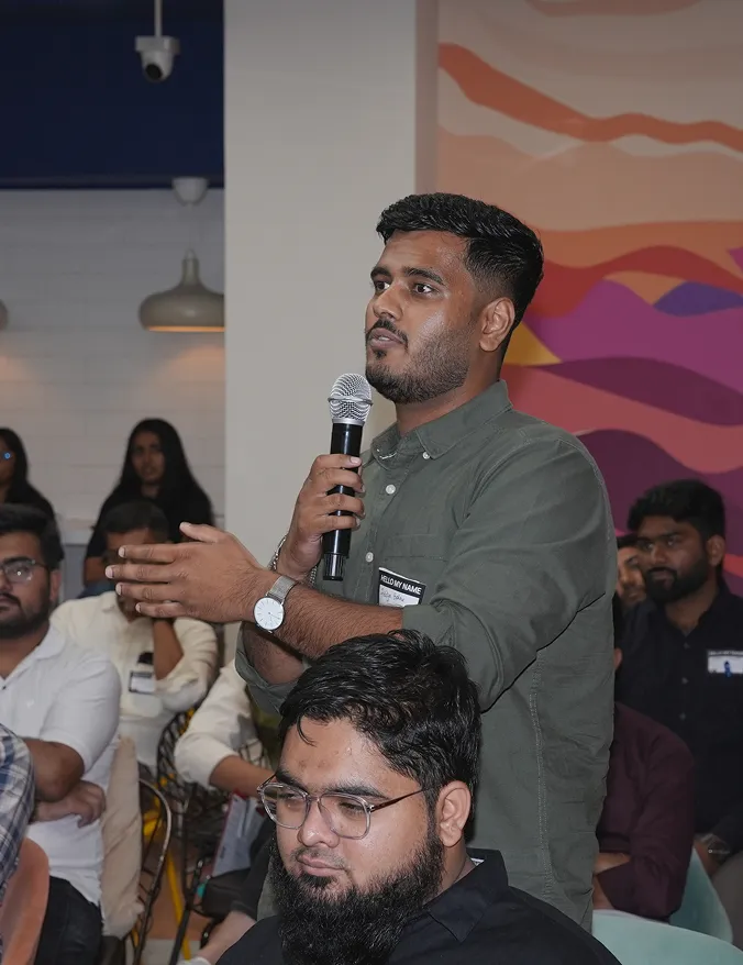 Man in green shirt speaking into microphone during a seated group event with colorful wall art.