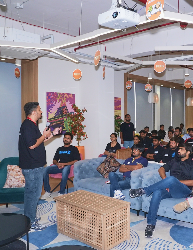 Presenter speaking to an audience seated on sofas and chairs in a modern room with hanging signs.