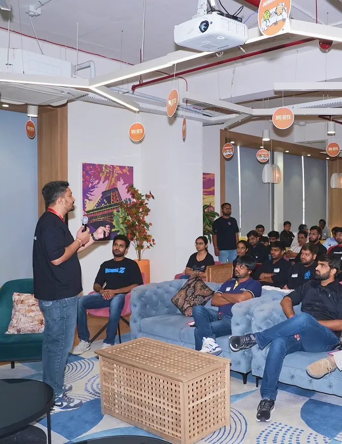 Presenter speaking to an audience seated on sofas and chairs in a modern room with hanging signs.