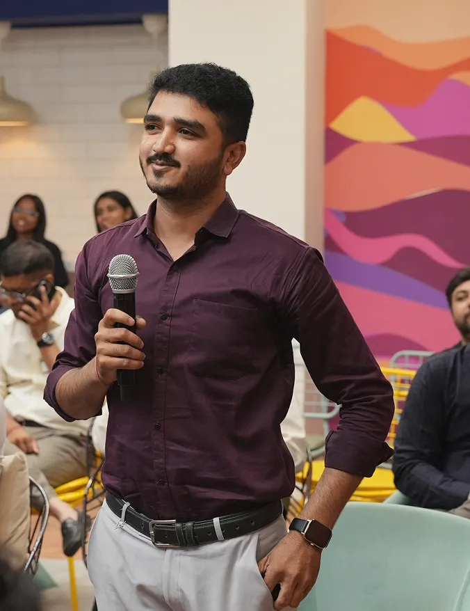 A man in a purple shirt holding a microphone, standing in a room with seated people and colorful wall art.