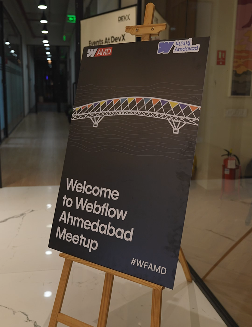 Sign on an easel welcoming attendees to Webflow Ahmedabad Meetup with a colorful bridge design and #WFAMD hashtag.