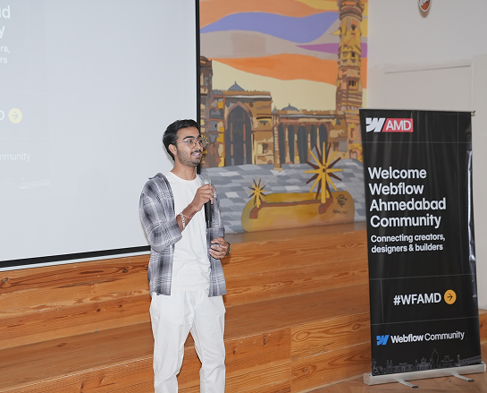 A man speaking on stage with microphone beside Webflow Ahmedabad Community event banner.