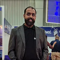 Man with beard wearing a gray blazer and black shirt standing indoors at a public event.