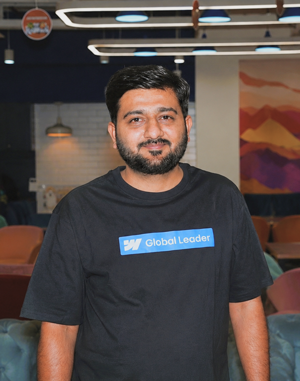 Man with short hair and beard wearing a black shirt that says Global Leader standing indoors.
