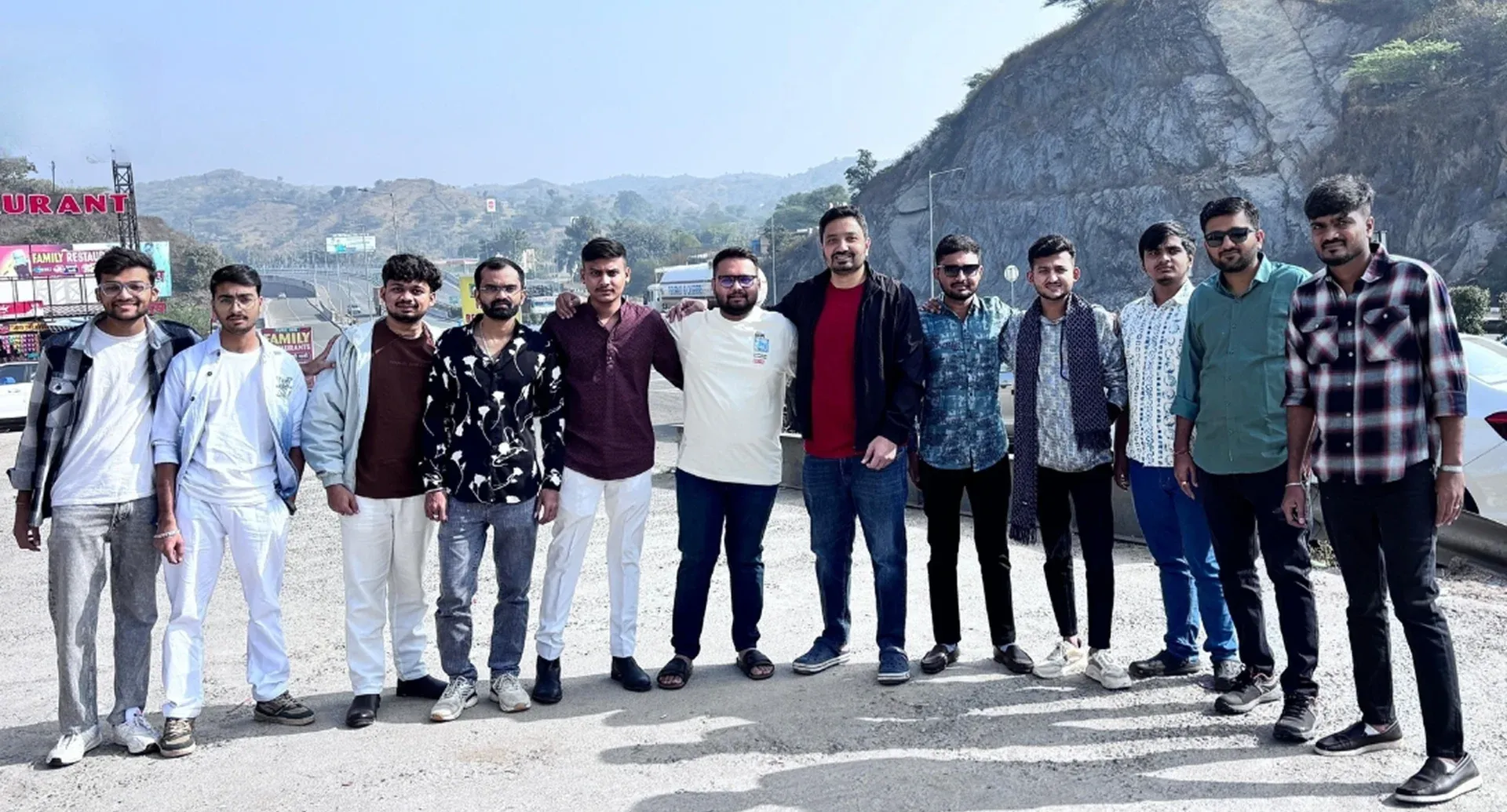 Group of twelve men standing together outdoors with mountains and road in the background.