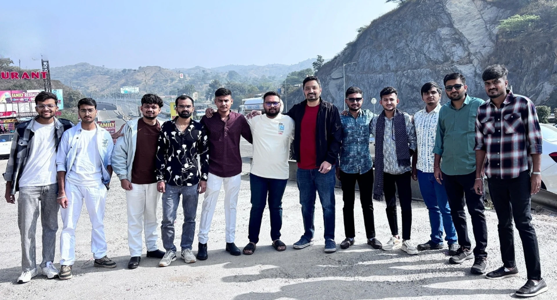 Group of twelve men standing together outdoors with mountains and road in the background.