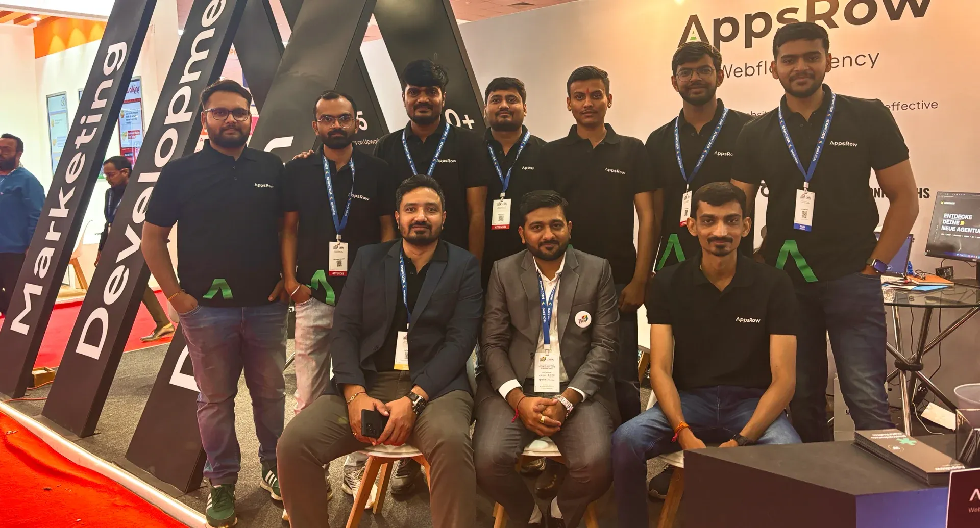 Group of nine men posing at a conference booth with Appsrow branding and badges.