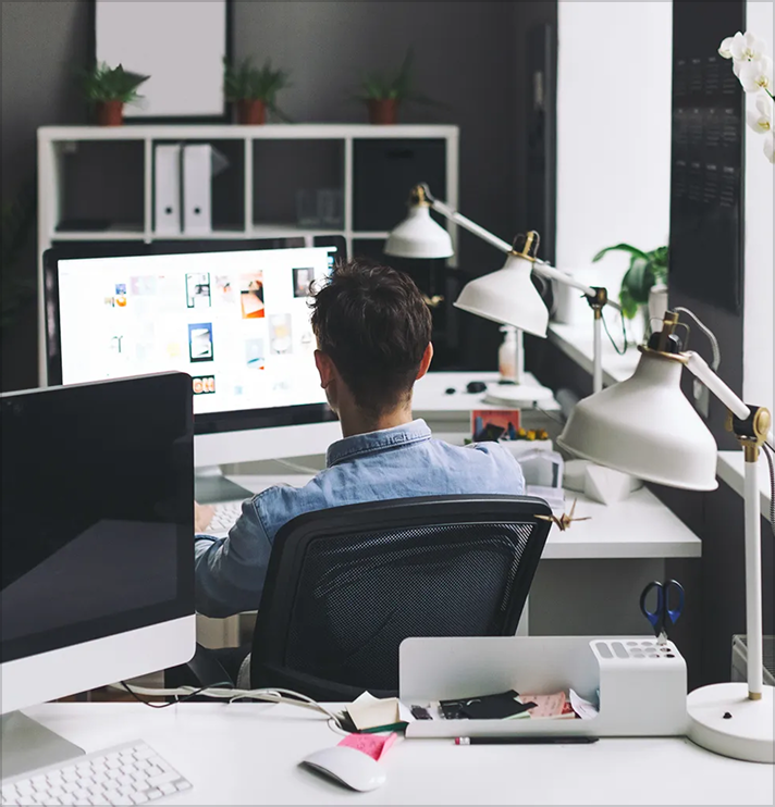 Person seated at a desk working on a computer with a bright screen in a modern office space featuring desk lamps, plants, and office supplies.