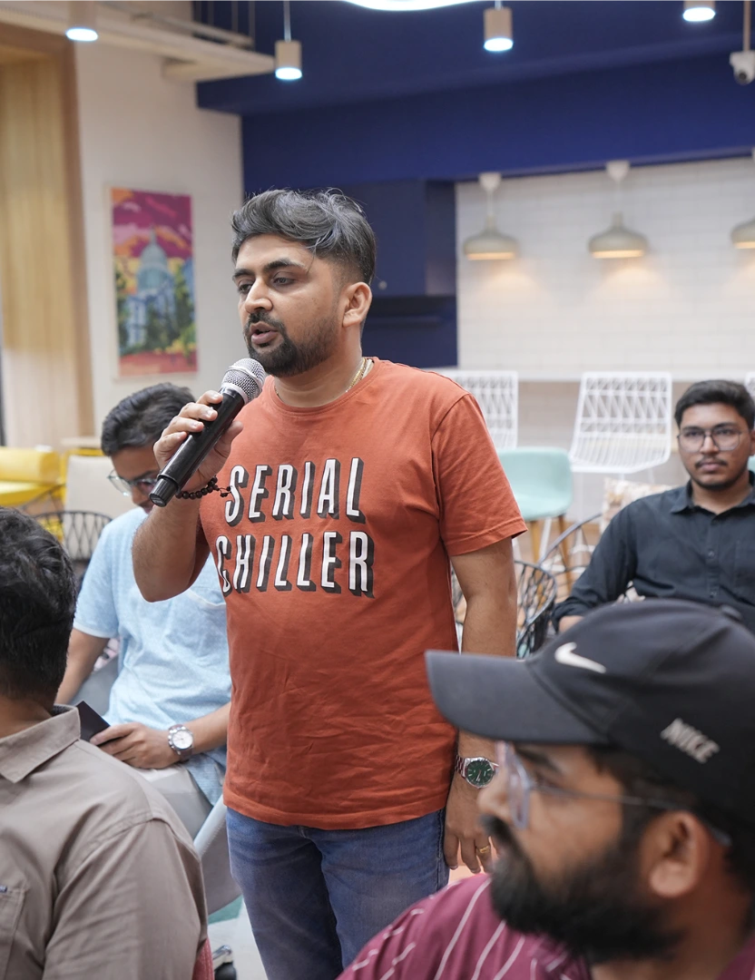 Man in orange 'Serial Chiller' shirt speaking into a microphone at an indoor event with seated audience.