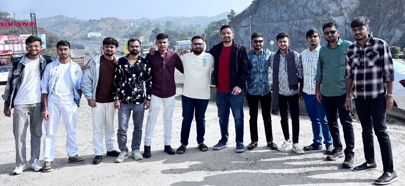 Group of twelve young men standing side by side on a sunny roadside with hills in the background.