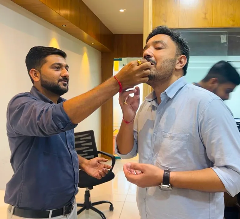 Man feeding another man a sweet in an office setting with outgoing man in background.