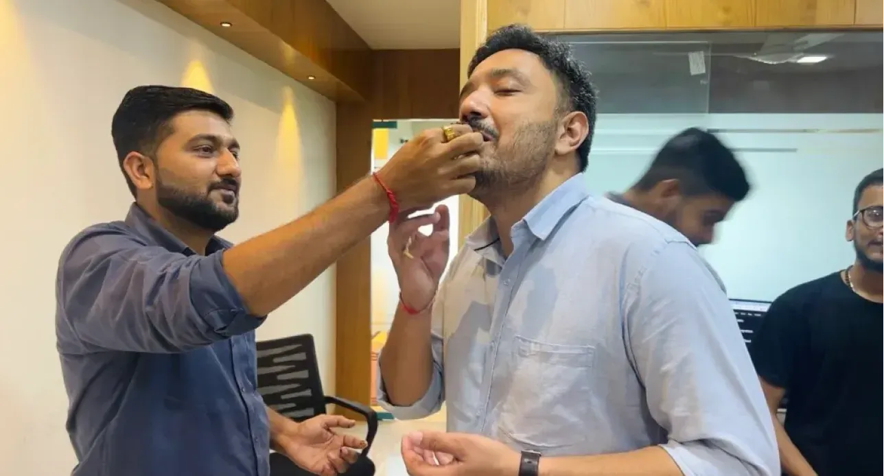 Man in blue shirt feeding cake to another man in light blue shirt inside an office setting.