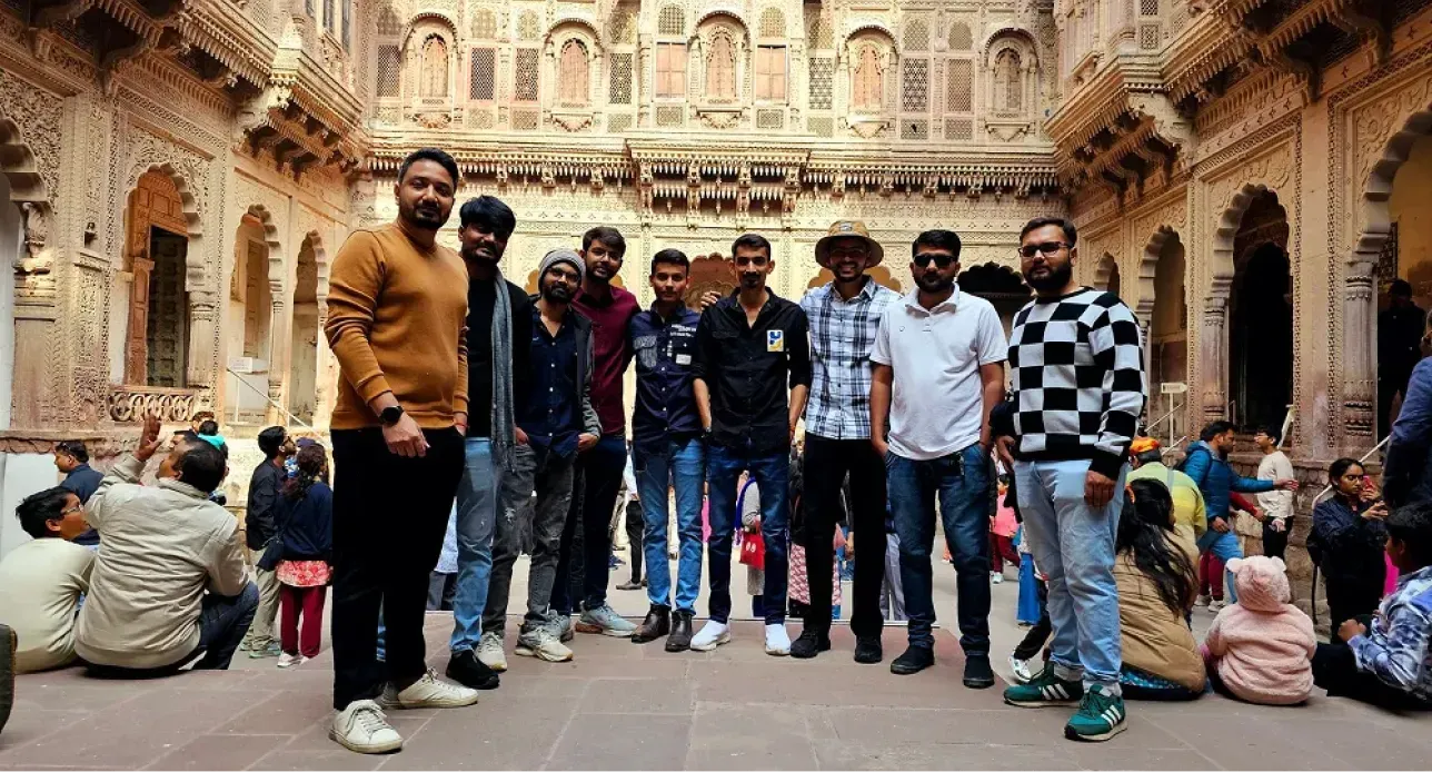 Group of men posing inside a historic ornately carved stone courtyard with visitors sitting nearby.