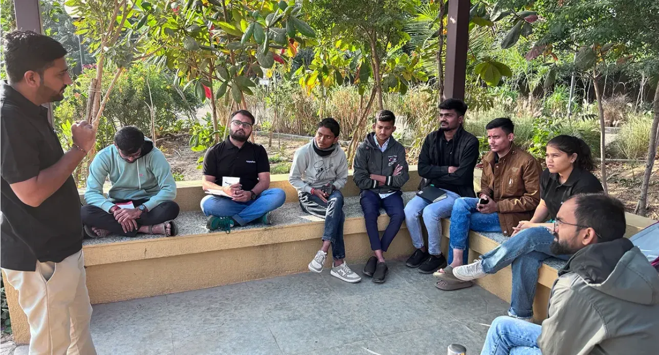 Group of people sitting on a curved bench outdoors while one man stands speaking to them.