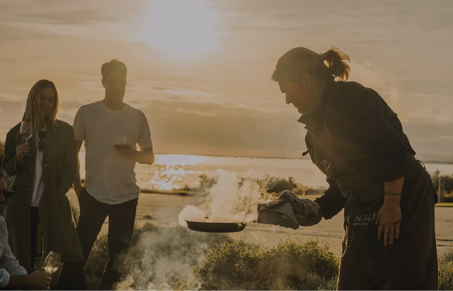 Person cooking outdoors over an open fire while three people watch holding wine glasses at sunset.