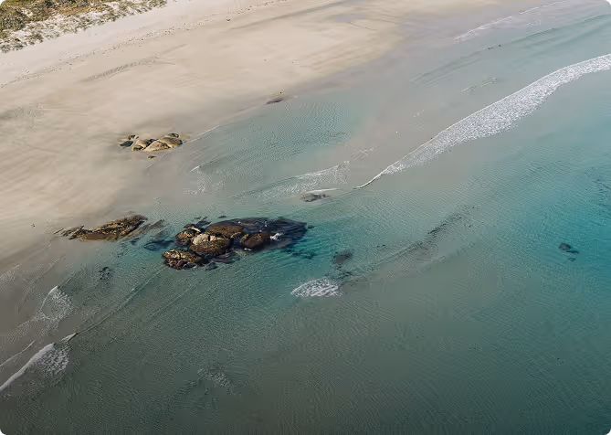 Aerial view of a sandy beach with shallow turquoise waters and clustered rocks near the shore.