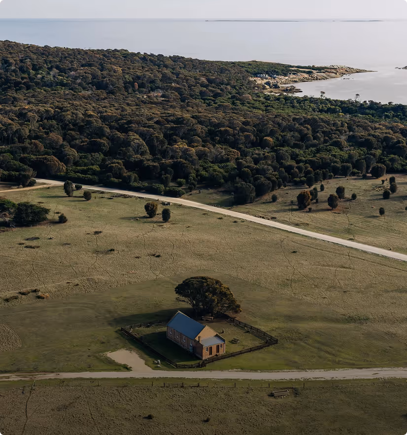 A small fenced house with a blue roof on open land near forest and water.