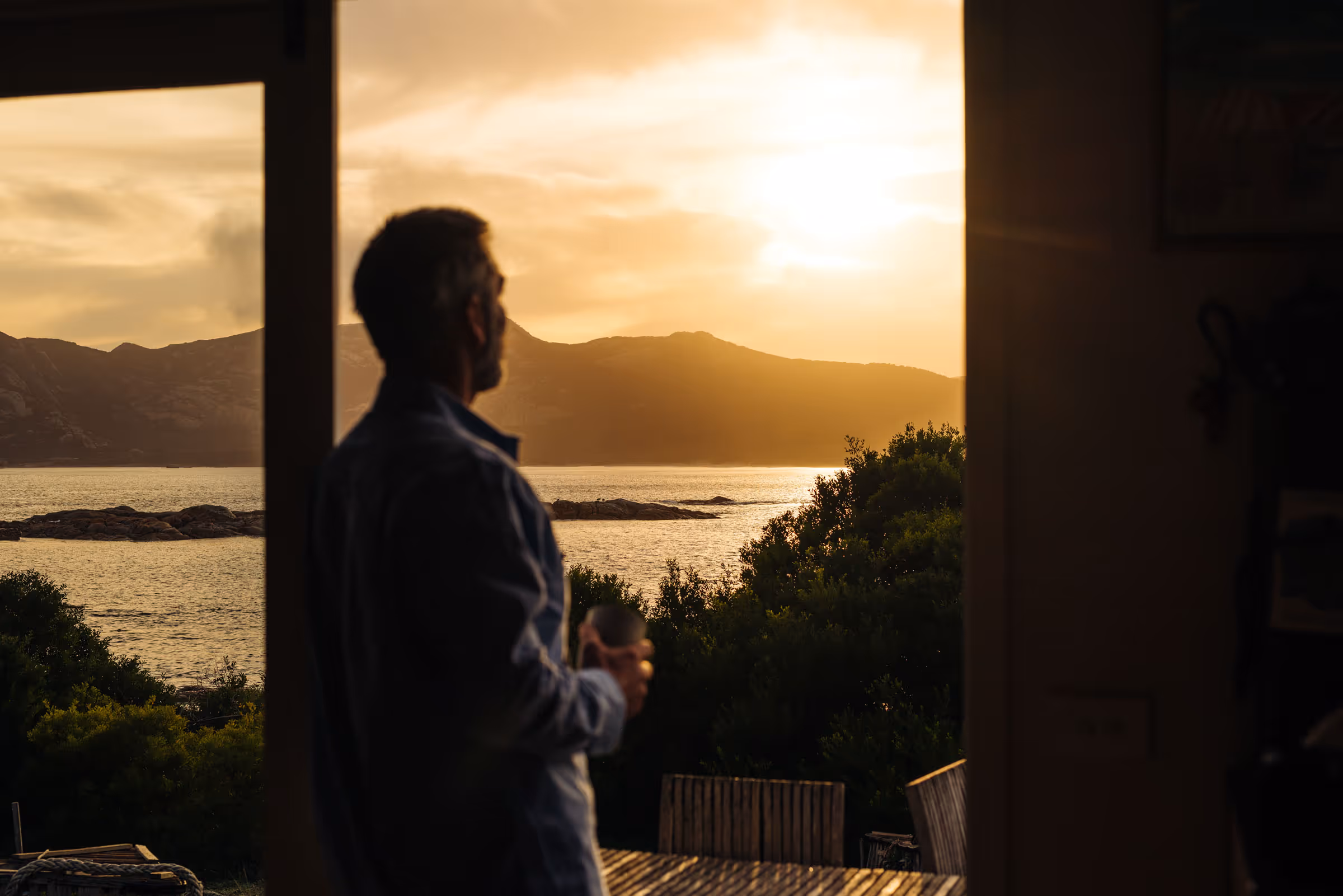 Man holding a drink indoors looking out at sunset over water, mountains, and trees outside.