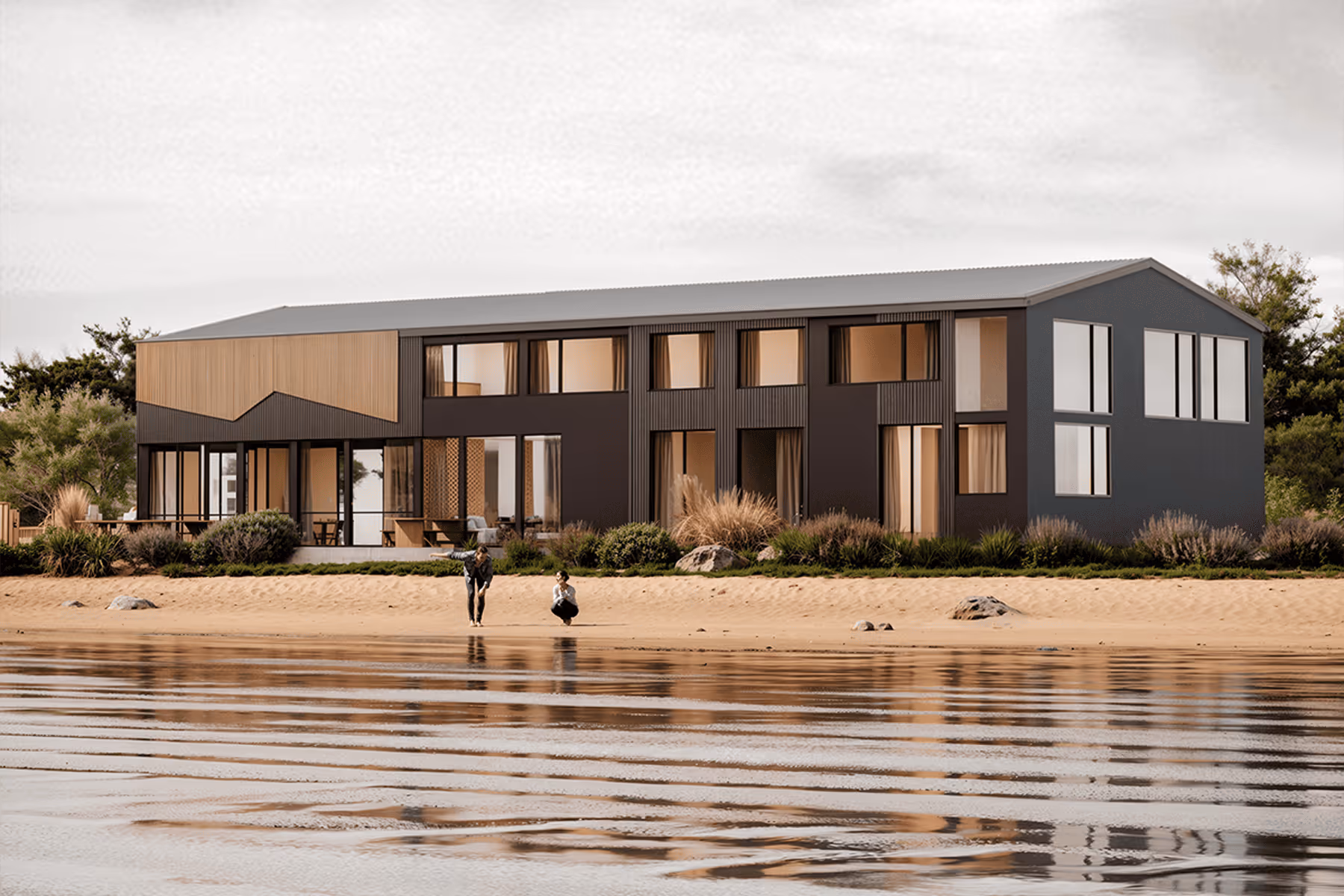 Modern two-story house with large windows by a sandy shore and calm water reflecting the building.