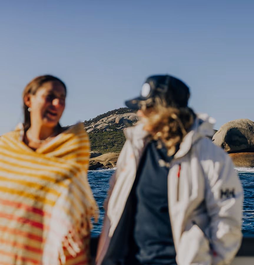 Two people talking outdoors near water with rocky hills and clear sky in the background.