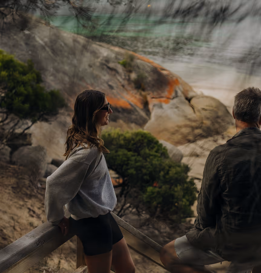 A woman and man relax on a wooden railing overlooking rocky terrain and distant water.