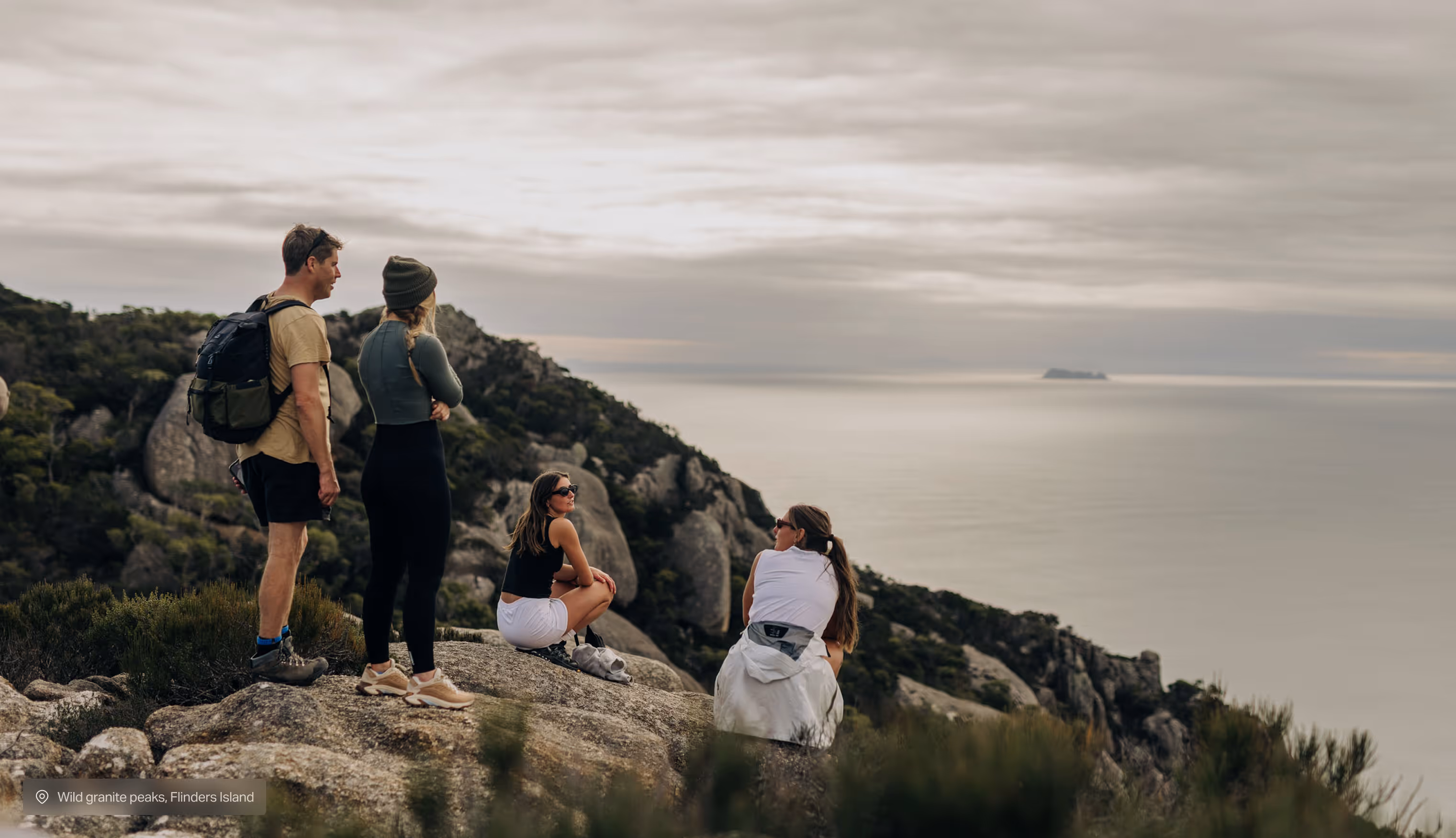 Two women sit and two people stand on rocky hill overlooking a calm sea and distant island.