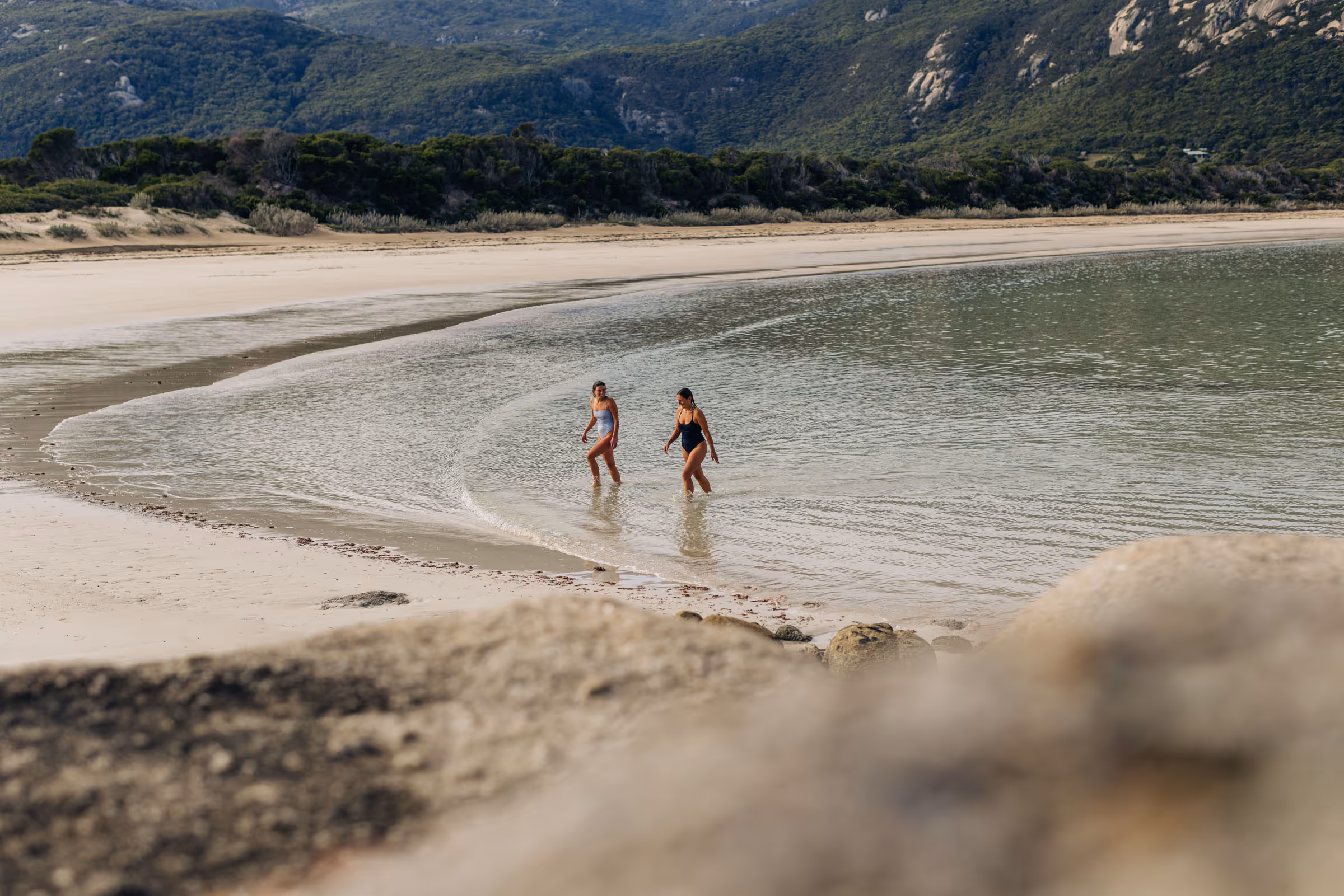 Two people in swimsuits walking in shallow water near a sandy beach with forested hills behind.