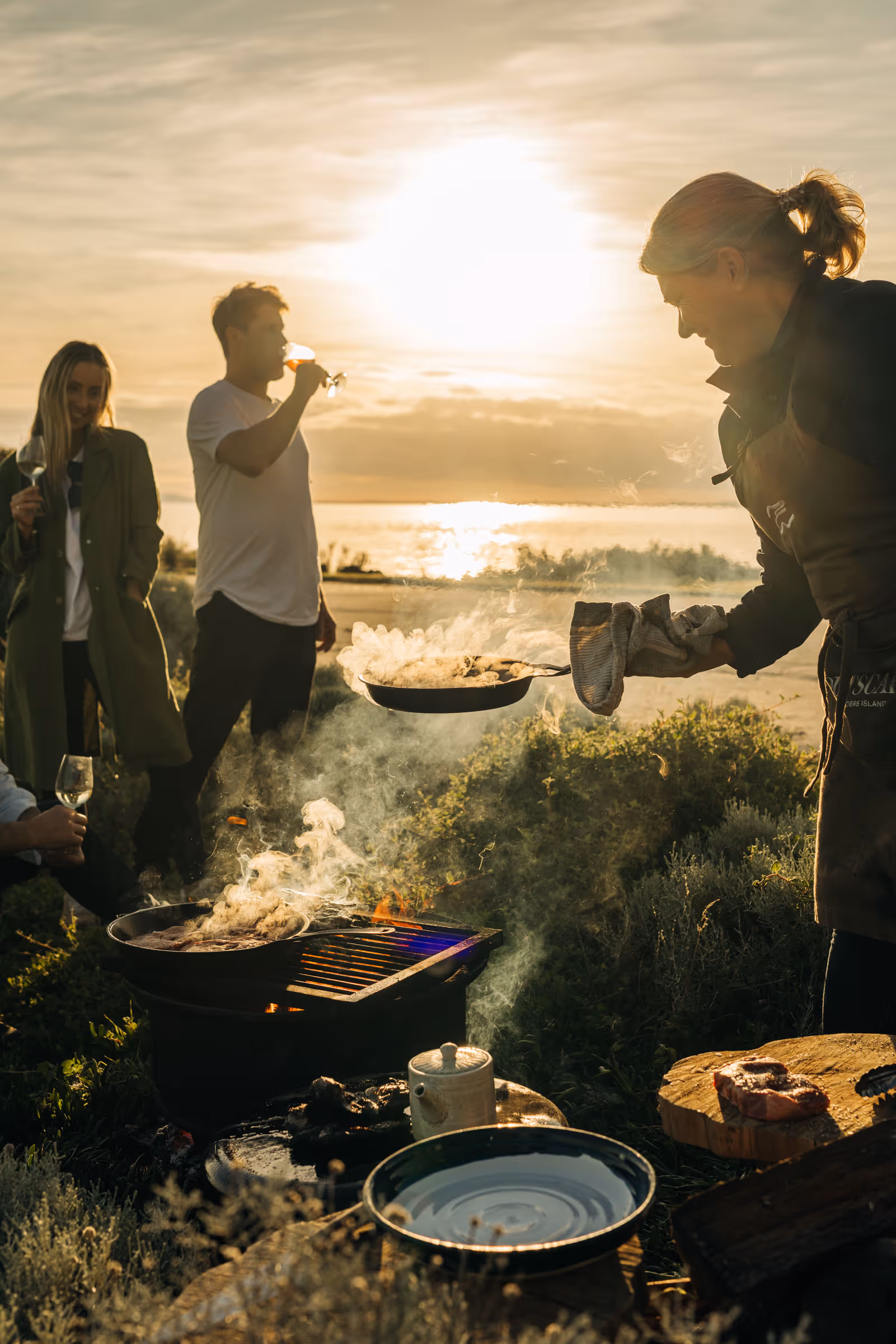 Person cooking over an outdoor grill with three people drinking wine at sunset by the water.