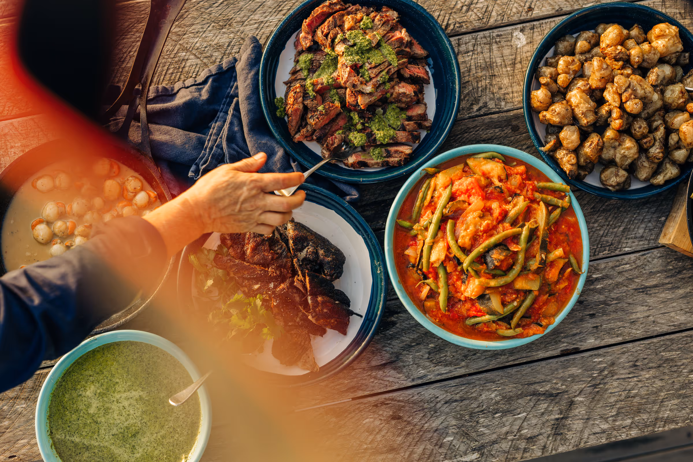 Hand serving sliced steak with green sauce among bowls of vegetables, cooked roots, and green sauce on wood table.