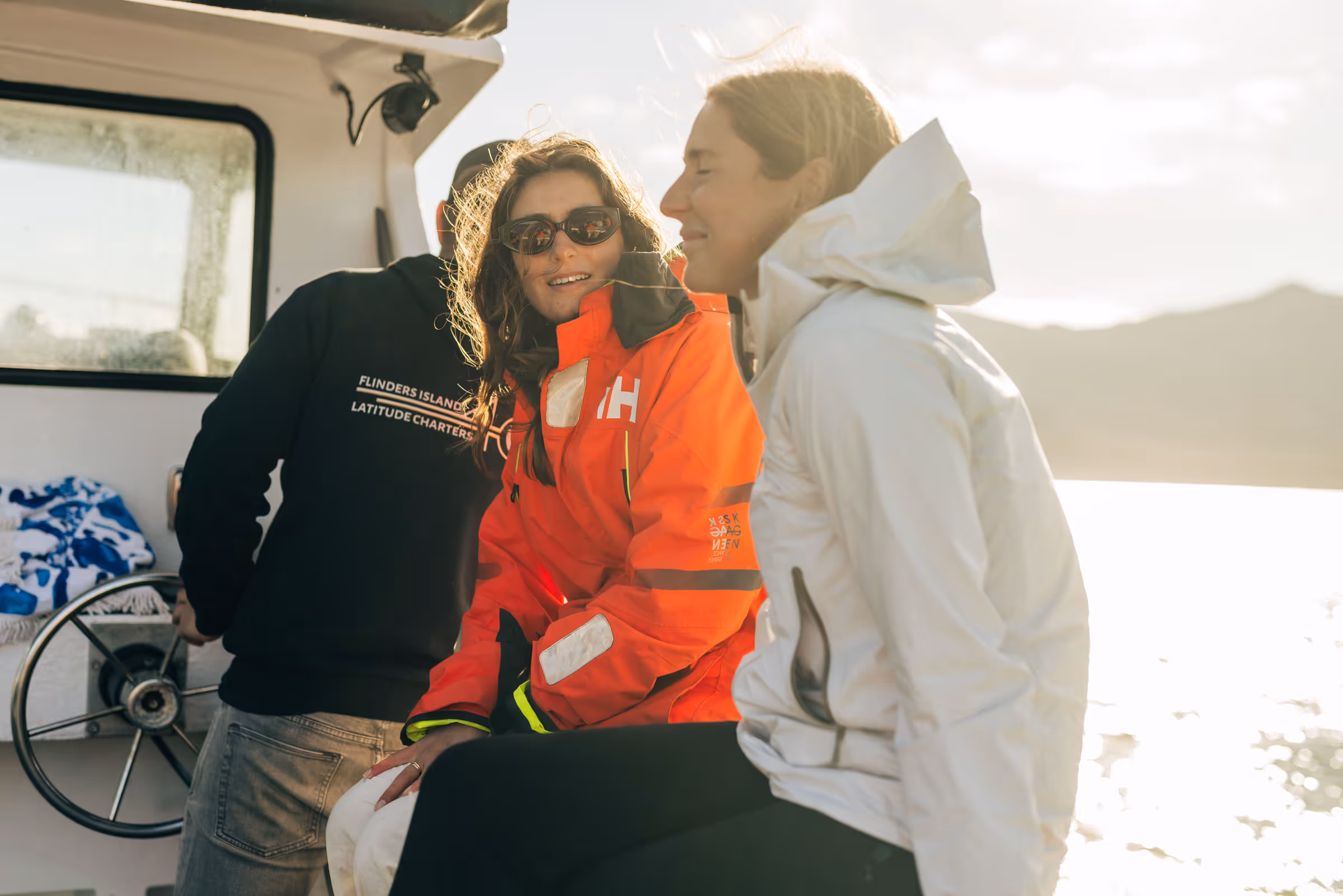 Three people wearing jackets on a boat during sunset with water and hills in the background.