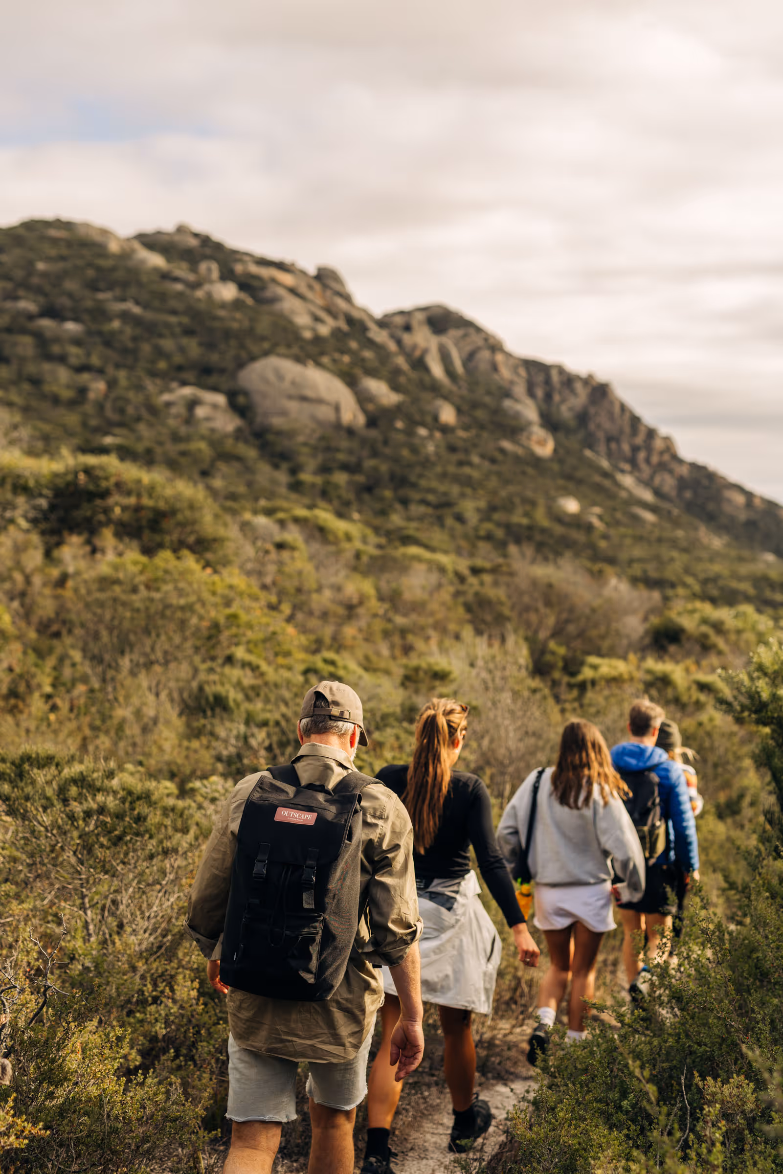 Group of hikers walking on a narrow trail through green shrubs with rocky hills in the background.