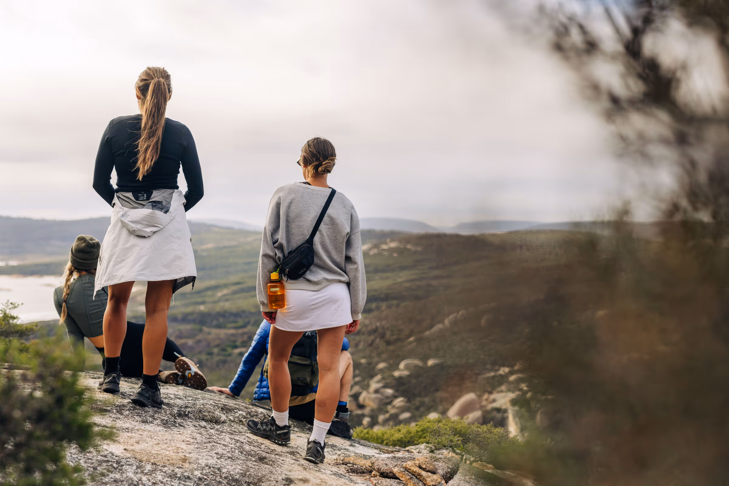 Four people on a rocky overlook wearing casual outdoor clothing, facing a vast mountainous landscape.