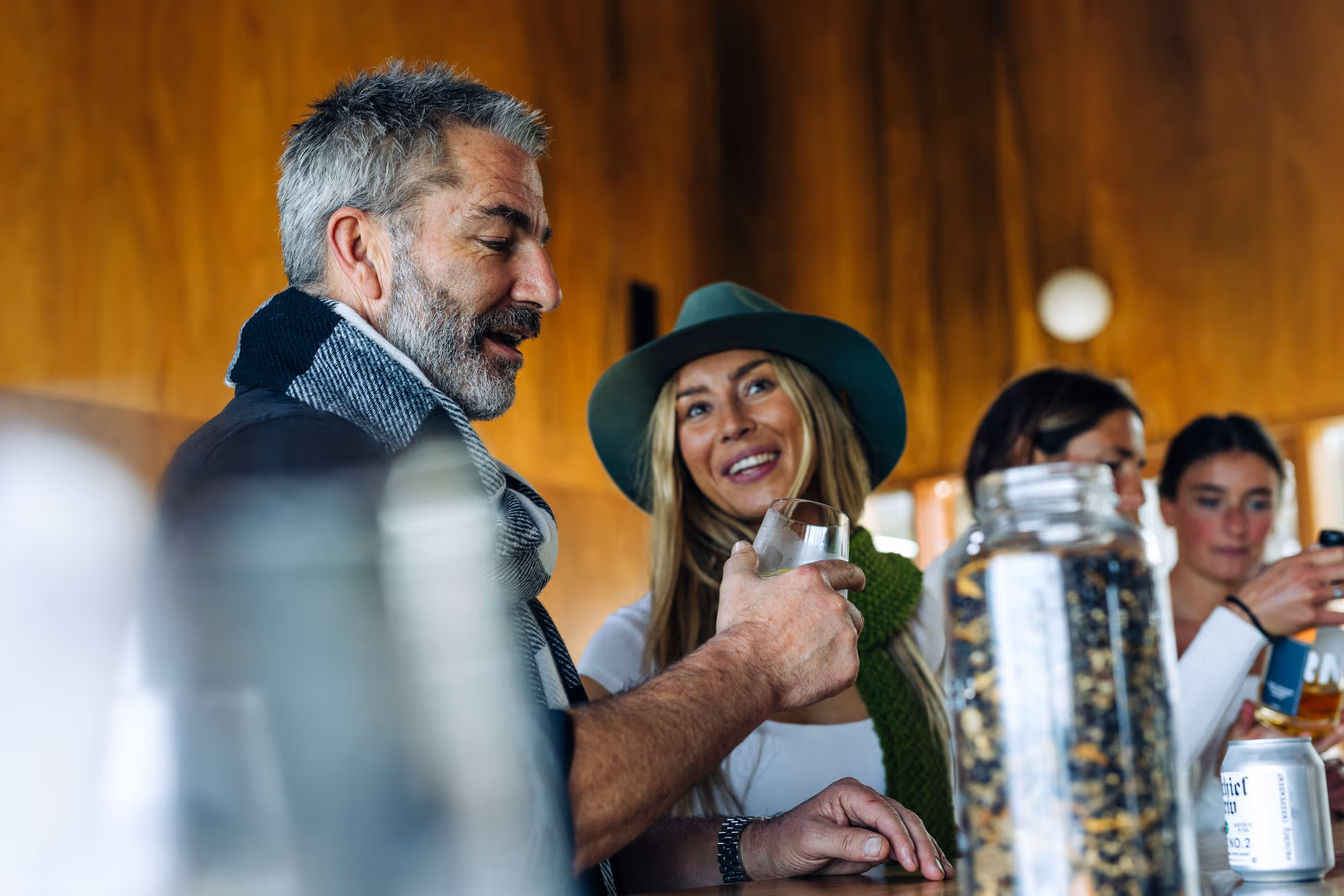 A man and woman smiling and holding drinks at a wooden bar counter with other people in background.