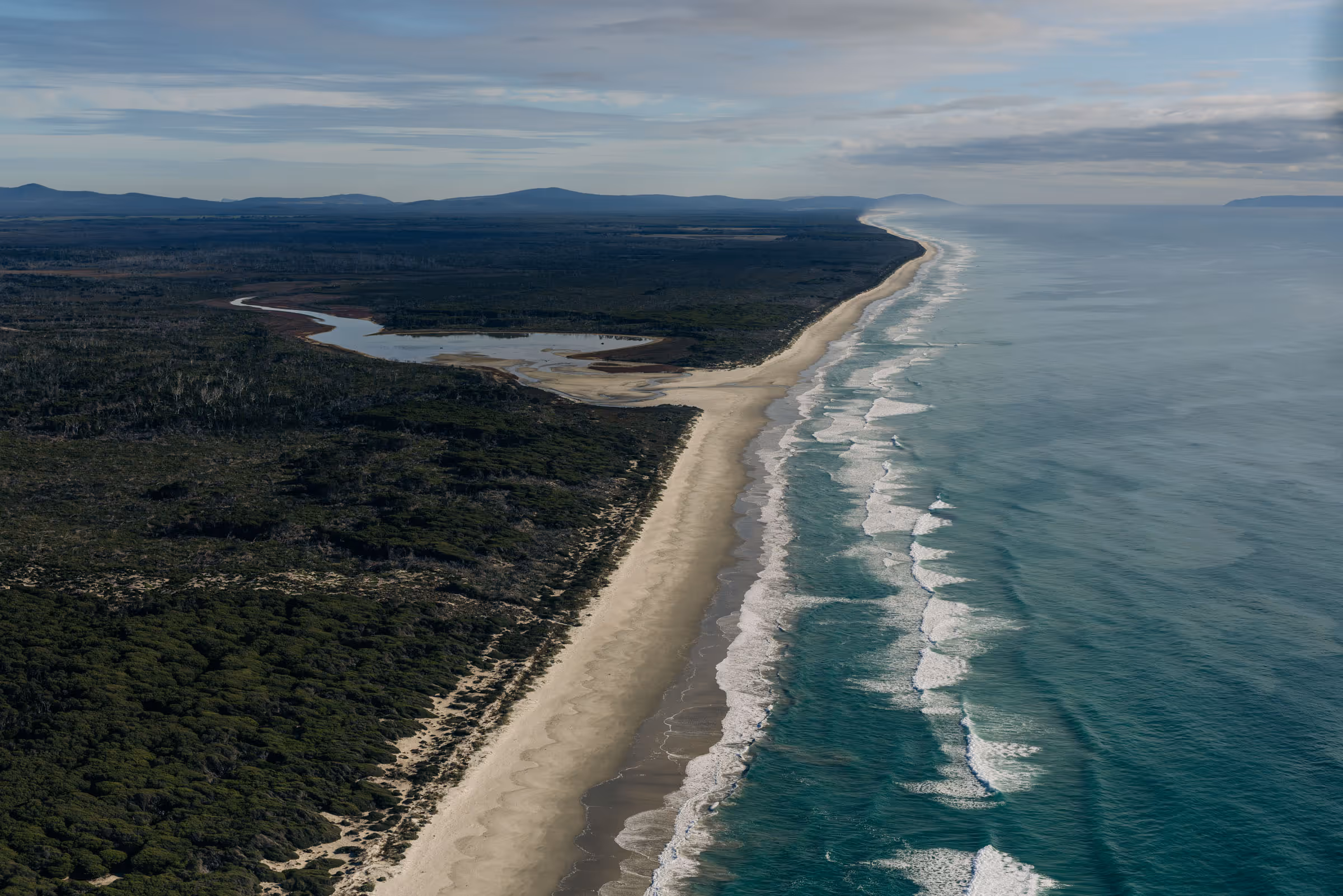 Aerial view of a long sandy beach bordered by forest and ocean waves under a partly cloudy sky.