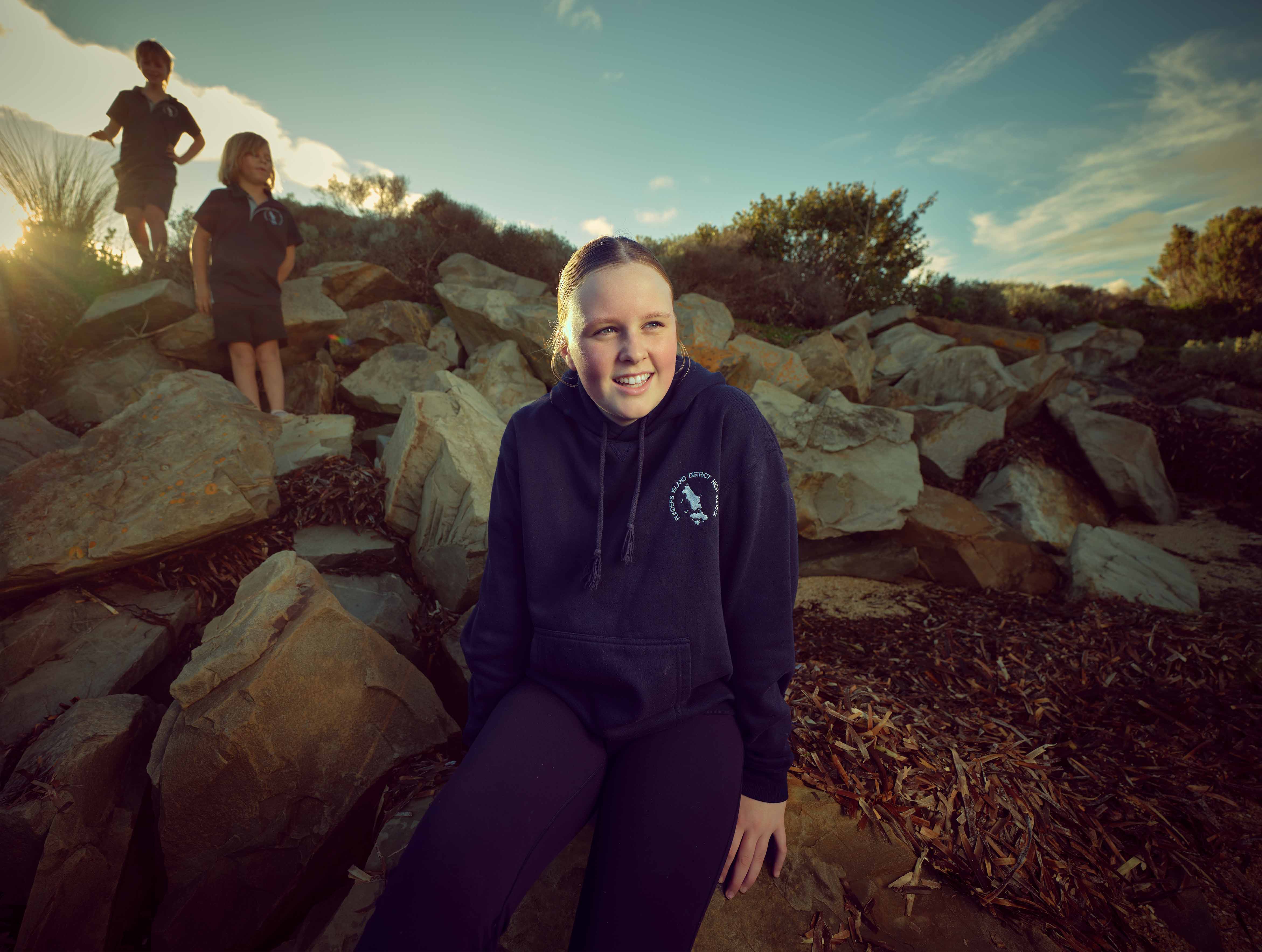 Teen girl in dark hoodie sitting on rocks smiling with two children standing on rocks behind her at sunset.