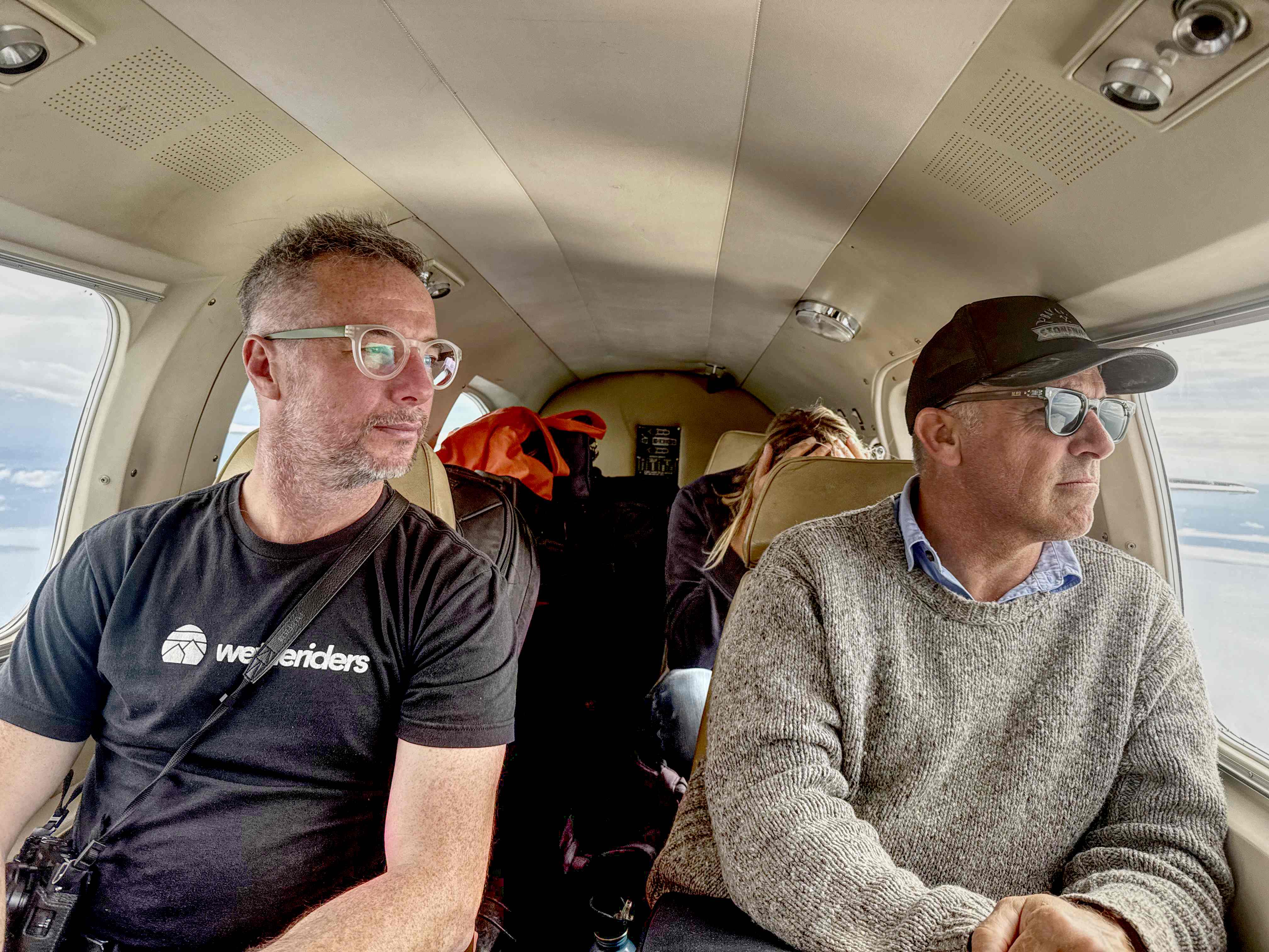 Three people seated inside a small plane looking out the windows, with bags in the back cabin.