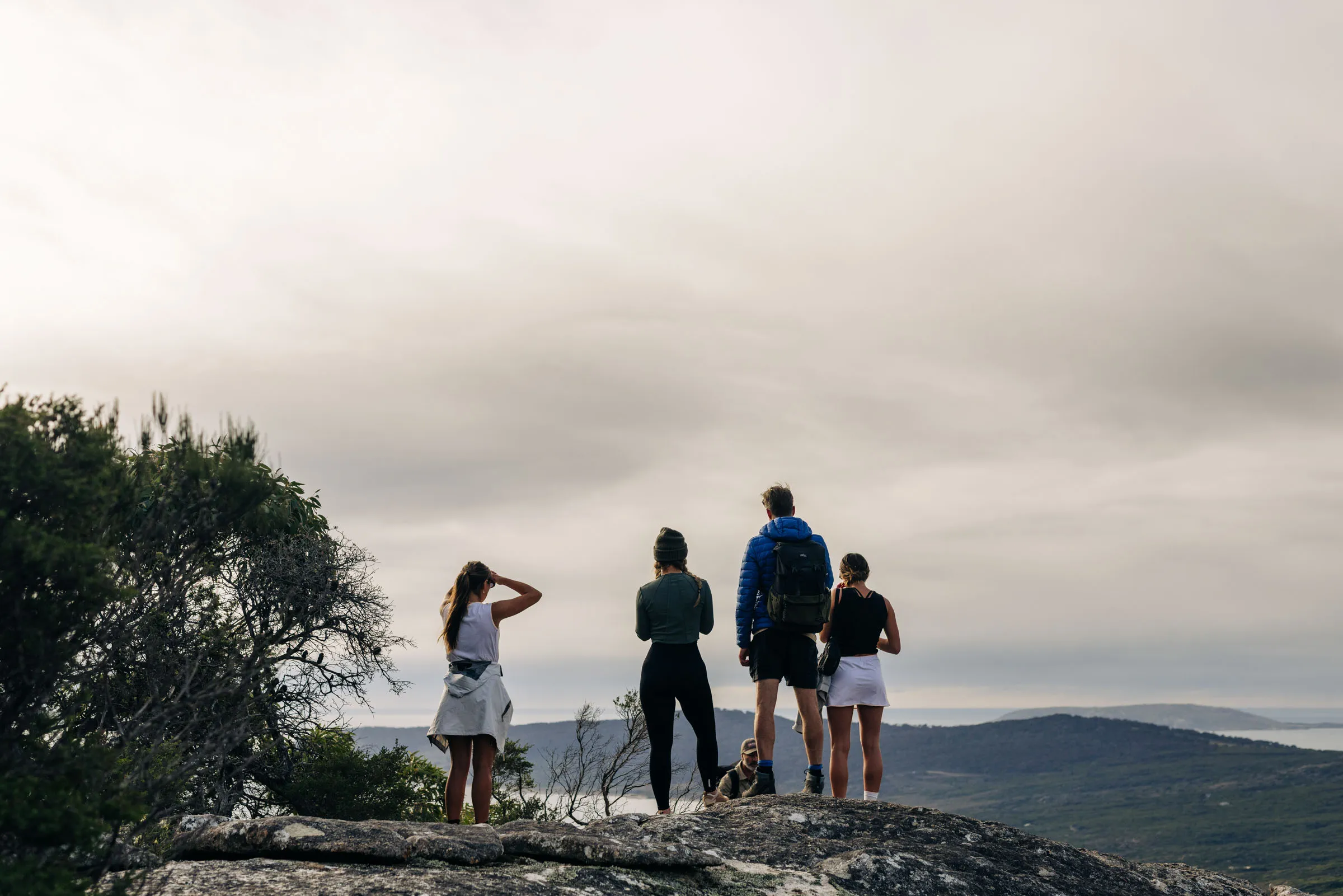 Four people standing on rocky hilltop overlooking hilly landscape under cloudy sky.