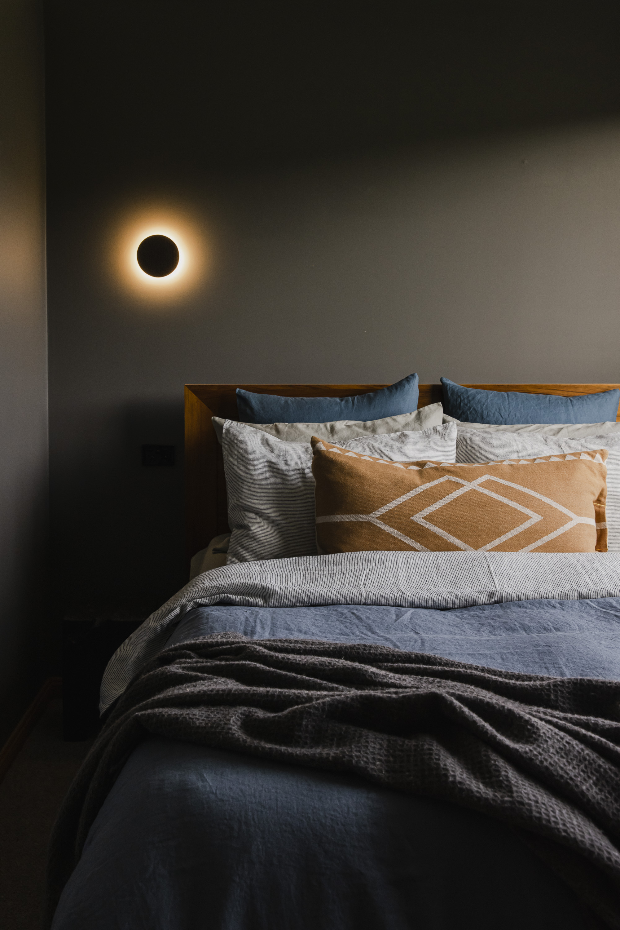 Bed with blue and beige pillows and blankets beneath a modern circular wall light on gray wall.