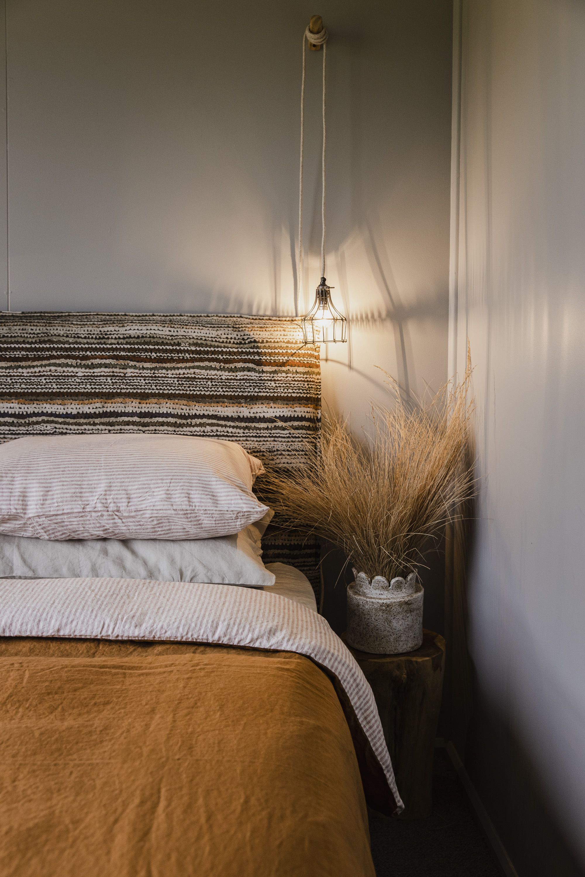 Bed with patterned headboard, soft pillows, orange blanket, a lit hanging lamp, and dried grasses in a vase.